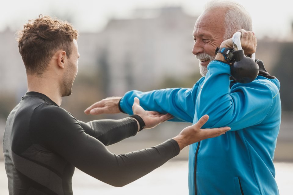 Huấn luyện viên chỉnh tư thế cho người đàn ông lớn tuổi tập ngoài trời — nghề health coach là gì